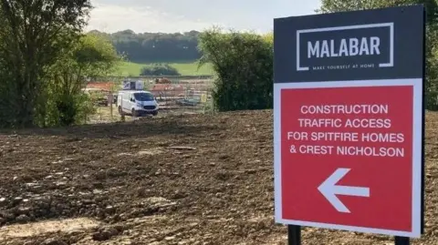 A red sign directing construction traffic to the Malabar estate. The project has only just started, so in the background is fencing and a van but no homes. The site is mostly muddy farmland, but it also has trees and bushes.