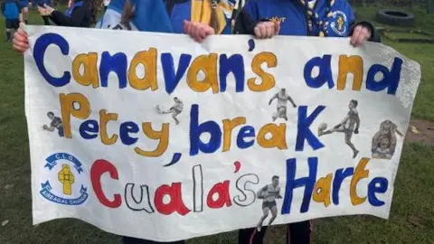 A homemade sign saying 'Canavan's and Petey, break Cuala's Harte' being held by two schoolchildren