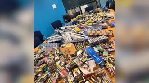 A table in a meeting room piled high with tobacco pouches, cigarette cartons and boxes of vapes.