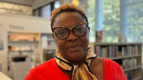 BBC Alexis Ekwueme - A woman with short hair wearing gold and tortoise shell rimmed glasses, a patterned scarf and a red top looks at the camera. She is standing in the middle of Ormeau Road library.
