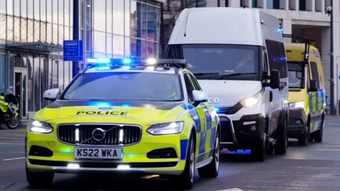 PA Media A white prison van drives between a fluorescent green and blue police car and a yellow police van on a road near Liverpool Crown Court
