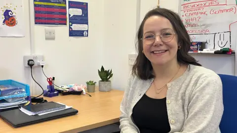 Break charity mentoring lead Maren Albrigtsen sits at a blue office chair by a wooden desk. She is wearing a grey cardigan and black top. She wears glasses and has brown hair cut in a bob. She is smiling at the camera. In the background there are notebooks and 2 pot plants on the desk. 