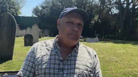 man wearing blue cap and checkered shirt sat in sunny cemetery.