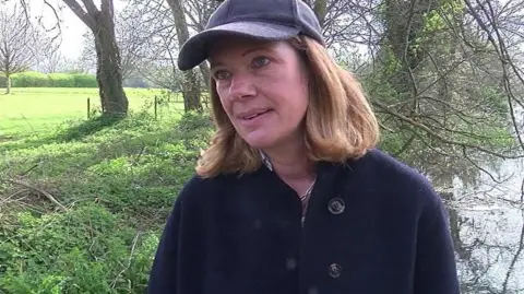 Claudie Paddick - she is wearing a navy coat and hat and is standing by a river.