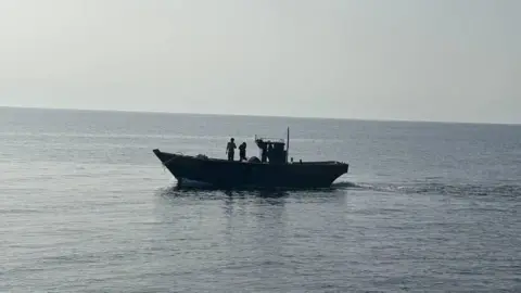 A wooden boat in the middle of the sea. Two people are seen standing on the boat
