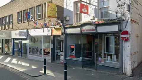 A branch of Santander in a white historic building with grey window frames is shown with other shops alongside. Two black bollards are in front of the building.