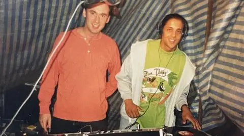 Chris Brown DJs Chris Brown (left) and Stu Banks are standing behind the decks at a rave. The image dates from 1989 and is grainy. Brown is wearing an orange top and Banks has a green T-shirt and white jacket on. It looks as though they are standing under a striped awning to protect them from the weather.