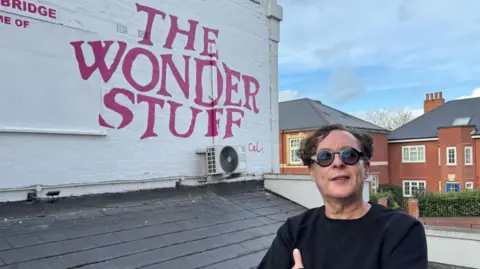 The Wonder Stuff lead singer Miles Hunt stands in front of a red logo which says "The Wonder Stuff" and also has Cal in red letters. The sign is on a white wall with a dark roof under it. Miles is wearing a black jumper and sunglasses and has his arms folded in front of him. 