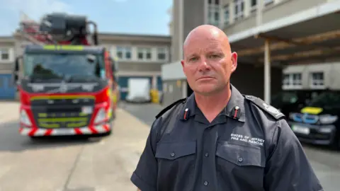 Bryn Coleman - a man wearing a black shirt fire uniform, behind is a fire engine on the left and parked cars to the right. Blue skies.