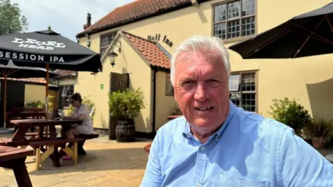Andrew Turner/BBC David Moss is wearing a blue shirt. He has white receding hair, and is sitting at a table outside The Bell Inn in Carlton Colville, of several pubs he operates. The building is rendered, and painted a ochre yellow. There are other tables nearby, with black parasols above them.
