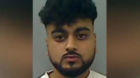 A mugshot of Hussain. He has black curly hair and a goatee and is staring into the camera.