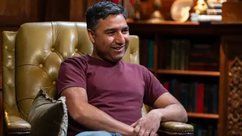Nick Mohammed is sitting in a large leather armchair. He is a youngish man with dark hair and a bit of stubble and wearing a dark red top with jeans.
