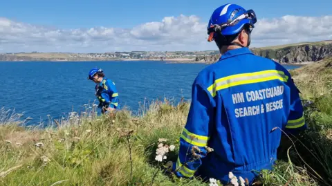 Two HM Coastguard search and rescue team members in blue uniforms and helmets stand on a grassy cliff overlooking the sea, appearing to carry out an operation along the coastline.