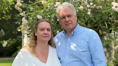Anthony Newborough wearing a light blue shirt with his arm round Elaine Newborough who is wearing a white top. They are in a garden.
