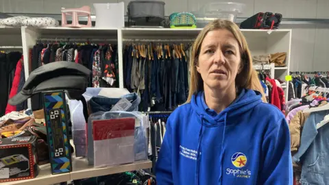 Stacey Southwell, a 42-year-old woman with long brown hair and wearing a blue hoody, is pictured standing in a charity shop with racks of clothes in the background.