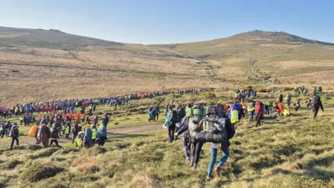 A view over the hills in Dartmoor. A big group of young people wearing hiking gear and carrying backpacks is spreading out over the area.
