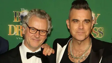 A man with grey hair and glasses and a black bow tie standing next to Robbie Williams