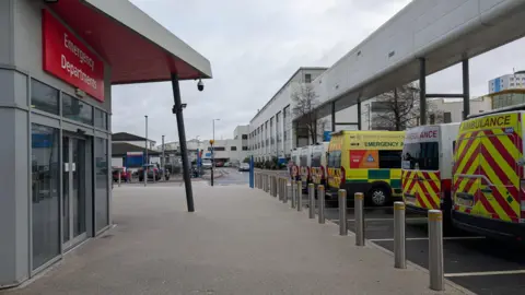 On the left is a building with automatic doors stating 'Emergency Departments'. Several ambulance vehicles are parked in front of it.