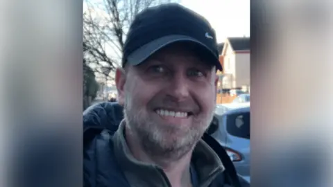 Adam Bee smiling at the camera. He has grey facial hair and is wearing a black cap with a small, silver Nike tick on one side. He is also wearing a grey, collared fleece and a black, padded, hooded coat. He is stood in the street, houses and cars can be seen in the background. 
