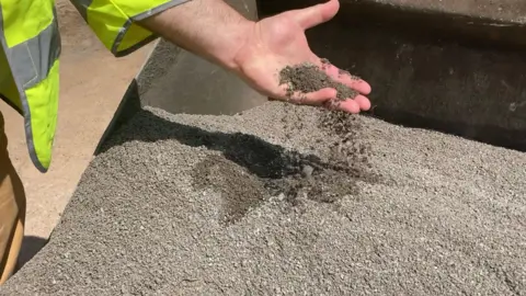 A hand pours small grey gravel from a container, with a high‑visibility sleeve visible.
