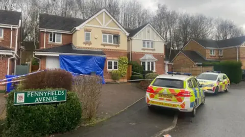 A detached house had blue tarpaulin over the white garage door and police tape sealing off the driveway. A police car is parked in front of the driveway and a fire service car is in front of it. There is a green street sign that reads Pennyfields Avenue. 