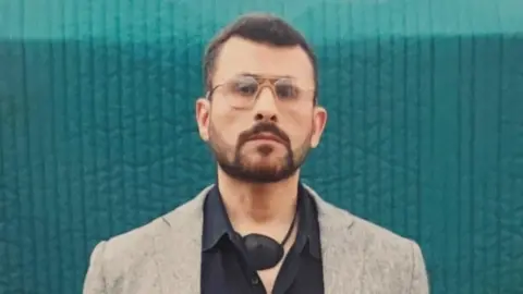 A head-to-chest shot of Abu Zubaydah, a man wearing glasses and with a defined beard, carrying an eye-patch around his neck. He is wearing a black shirt and grey blazer.