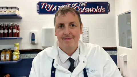 A photograph of a middle-aged man smiling at the camera inside a white-tiled shop. He is white, has brown eyes, brown hair and a moustache. He is wearing a white butchers coat emblazoned with "Lambournes" and a cow logo, standing in front of a large sign that reads: "Try a taste of the Cotswolds".