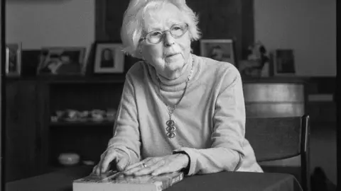 Mike Stone A black and white portrait of Agnes Kaposi wearing glasses and a necklace, resting her hands on a book whilst seated at a table.