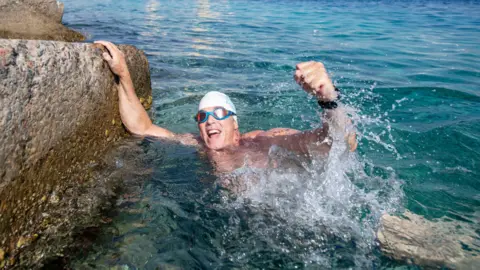 A smiling endurance swimmer wearing goggles and a swim cap raising a fist while clinging to a sea wall in clear blue water.