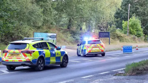 Two police cars block off a road, while a blue sign reads 'Police accident'  