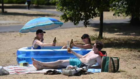 Andy Rain/EPA Two women sitting inside a children's paddling pool, both holding a bottle of alcohol. A man is lying on a towel, next to the paddling pool, holding a bottle of alcohol. They are at a park, and have bags besides them.