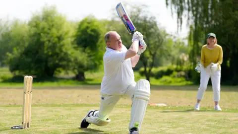 PA Media Sir Ed Davey lunges forward as he swings his cricket bat. A woman is in the background of the photo smiling and waiting to catch a ball.