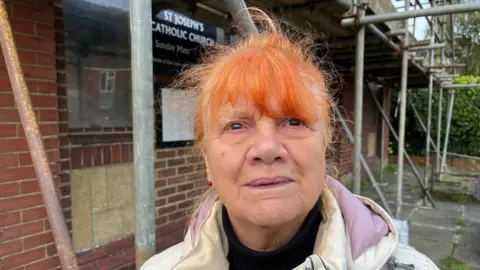 Phil Harrison/BBC Margaret Irving looks at the camera, behind her you can see the scaffolding on the outside of the building and the sign on the front of the church