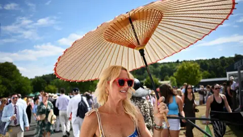 Reuters A woman with blonde hair and wearing a blue dress and red sunglasses smiling beneath a wicker umbrella on a sunny day, with crowds of people behind her.