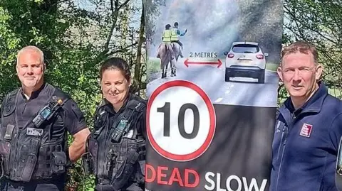 Two police officers and another man next to a sign that says "DEAD SLOW".