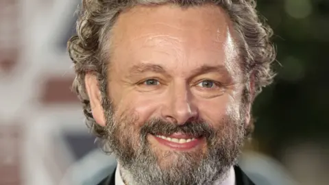 Getty Images A close up shot of Michael Sheen. He has a grey beard and curly greying hair, and smiles at the camera. The background is blurred. 
