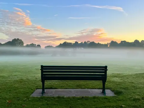 BBC Weather Watchers/Dammo A clearly defined layer of mist lies over grassland below a blue sky with the sun starting to rise behind trees. In the foreground, a traditional park bench sits on paving slabs.  