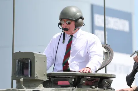 King Charles has the top half of his body sticking out of the top of a tank, he is wearing a white shirt with a red and green tie and some sunglasses. He is also wearing a green helmet with ear defenders and a microphone.