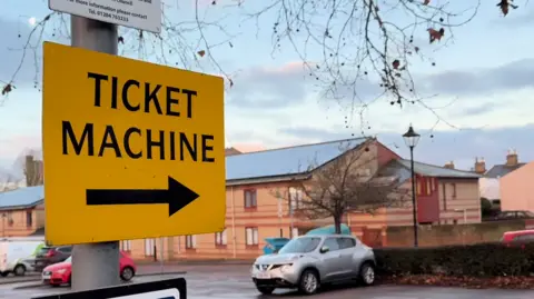 A car park with a yellow sign that reads ticket machine and a black arrow directing people toward it. Cars are parked in the car park.