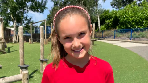 Beau is outside in the school grounds standing on grass and wearing a red t'shirt and her brown hair is tied back. She is smiling at the camera.