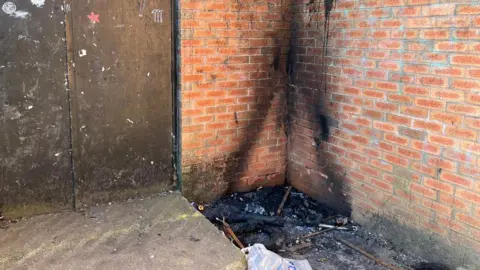 The burn marks of a fire which was lit next to the fire exit of a brick building. A pile of black soot and burnt waste can be seen next to a concrete step down from the fire exit.