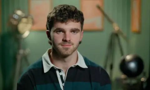A man, Tom, looks at the camera. He is wearing an blue and green rugby shirt