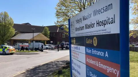 Exterior of Wrexham Maelor hospital showing a metal sign and vehicles outside the main A&E department