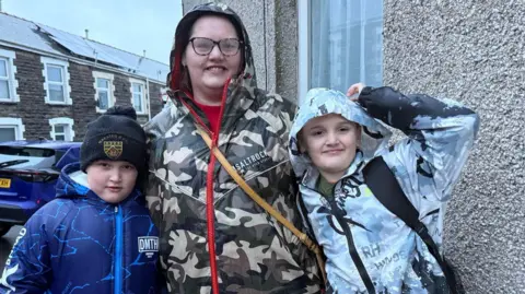 Bethan, wears a camouflage long rain coat with her hood up and glasses. Her two sons also wear raincoats with their hoods up and are pictured outside their terraced house