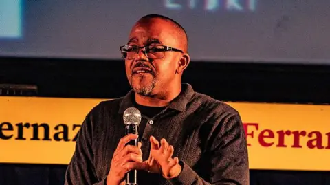 Getty Images Kehinde Andrews, a black man with a dark shirt, square-framed glasses and a black and grey beard, stands on stage holding a microphone.