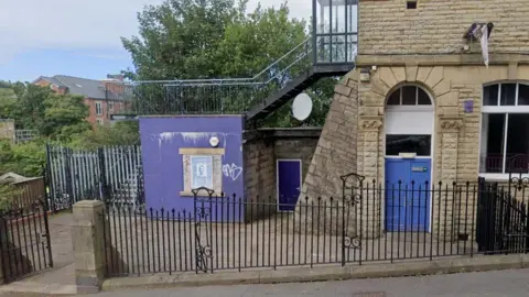 Google Street view of the outbuilding, a purple-painted square building at the side of the library surrounded by metal fencing