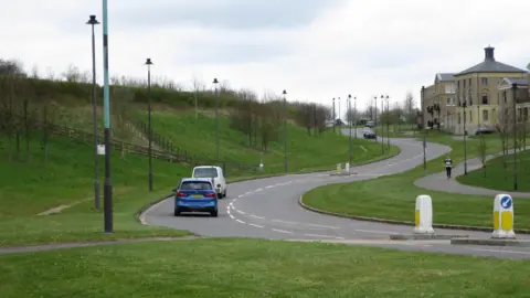 LDRS Two cars drive along Middle Farm Way as it snakes up a small incline on Poundbury's northern perimeter. The wide, two-way single carriageway has wide grass verges and some houses set back from the road on the right-hand side.