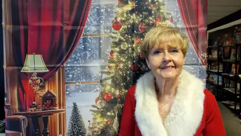 A woman with short blonde hair wearing a Santa-style coat stands in front of a backdrop depicting a Christmas tree.