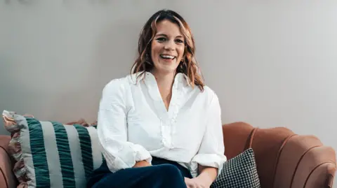 The ADHD Nurse A woman with shoulder length brown hair is wearing a white frilled shirt, and sitting on an orange-coloured sofa.