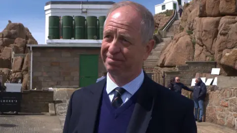 Ian Gallichan is smiling as he stands outside. He is wearing a navy jacket, navy jumper, blue and white striped shirt and a navy striped tie. Behind him are large boulders which form a wall and there are steps leading up through them. There is a small building behind him which has a door.